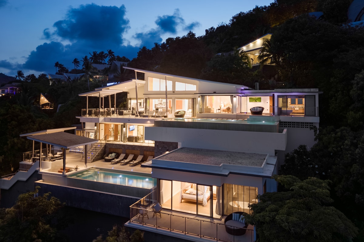 The modern villa is set against a twilight backdrop, showcasing expansive terraces and illuminated living spaces. Two infinity pools glisten under the evening sky, with lounge chairs arranged invitingly nearby. Large glass doors open to reveal indoor areas, maintaining a connection with the enchanting exterior.