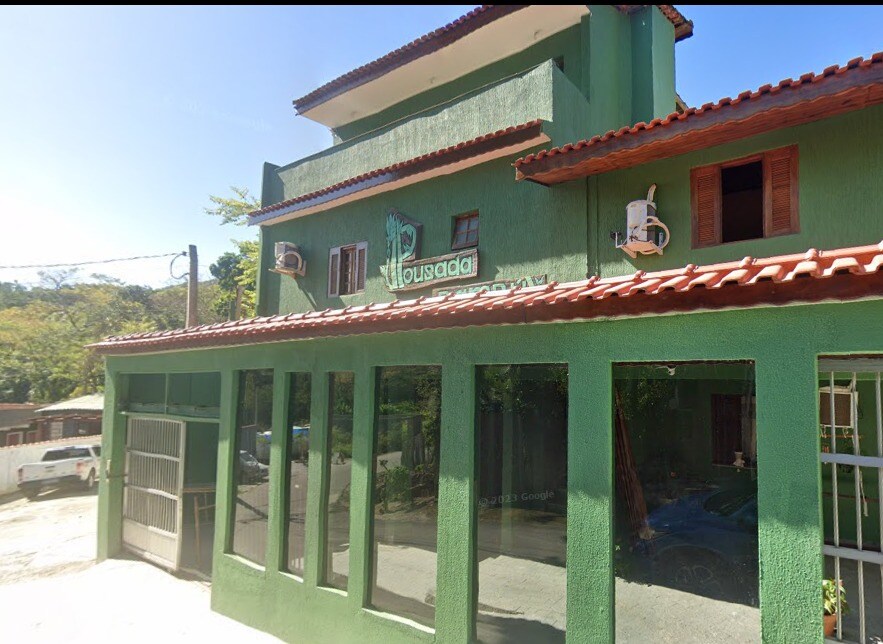 A green, three-story building is observed, featuring a red-tiled roof and large glass windows that reflect the surrounding nature. Air conditioning units are mounted on either side of the entrance, which is flanked by a simple design. The environment appears quiet and inviting.