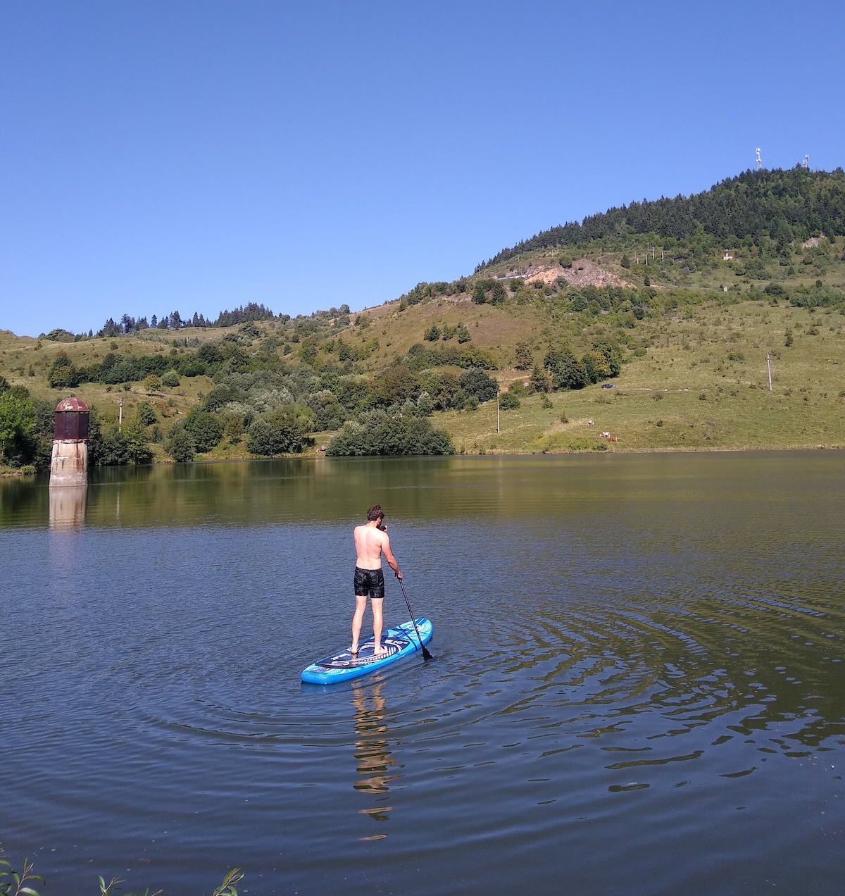 A person stands on a paddleboard, navigating calm waters. Lush green hills rise in the background, with trees dotting the landscape. A weathered structure is visible near the water's edge, blending into the serene scenery under a clear blue sky.