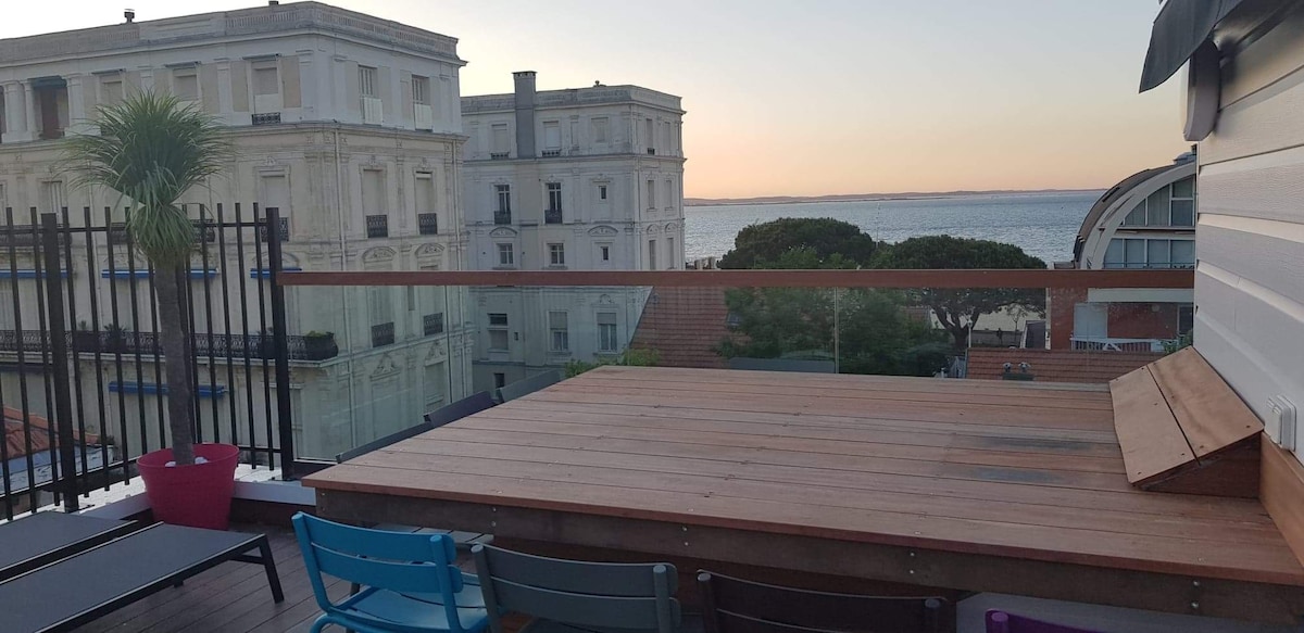 A rooftop terrace is showcased, featuring wooden decking and a cozy seating arrangement. Colorful chairs are positioned comfortably. The view encompasses nearby buildings and the sea, with a warm sunset casting a soft glow over the scene.
