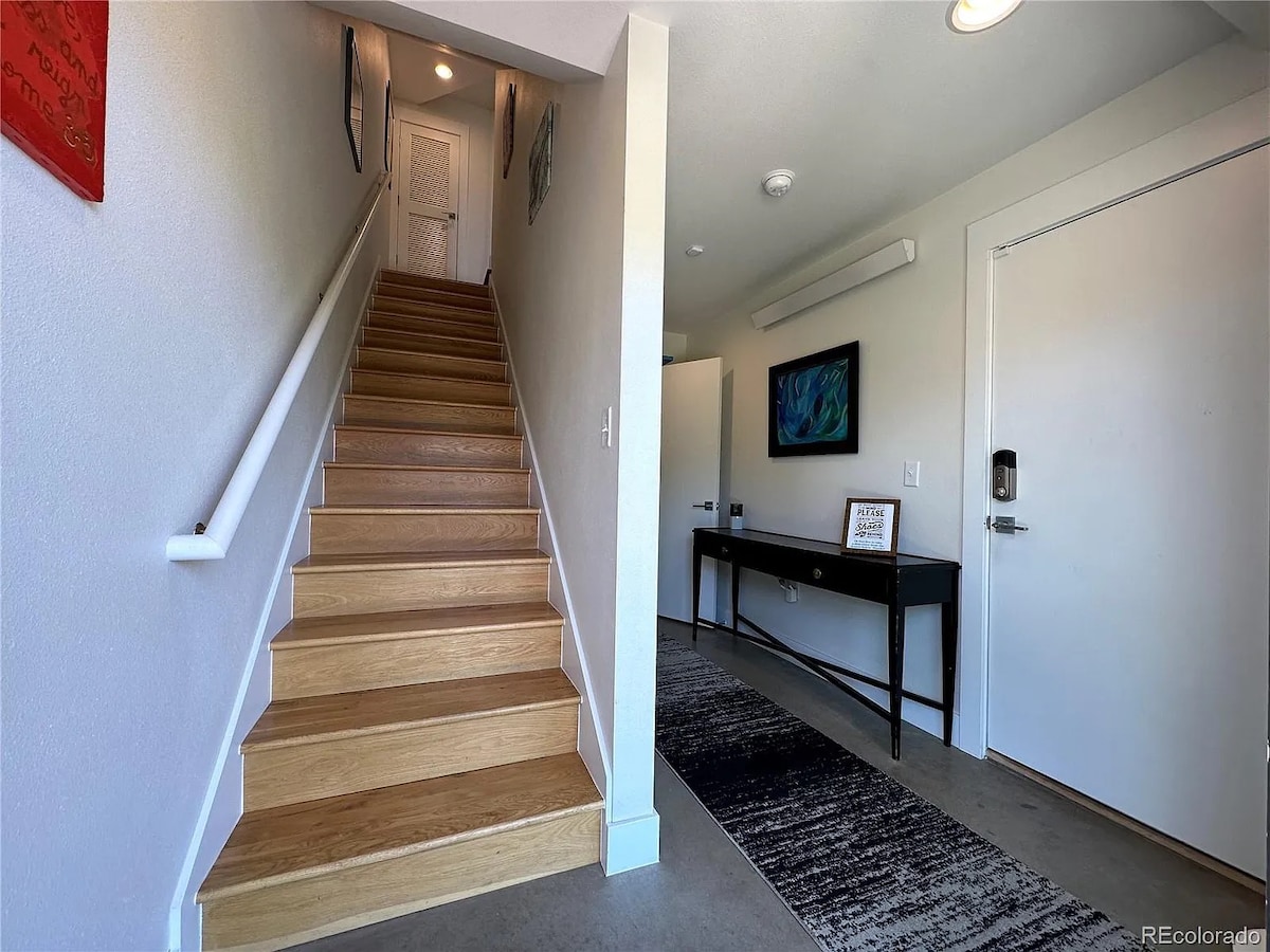 An entryway showcases a modern design with a staircase featuring polished wooden steps. A sleek console table is placed against the wall, complemented by a textured area rug. Soft lighting enhances the inviting atmosphere of the space, leading toward the open living area.