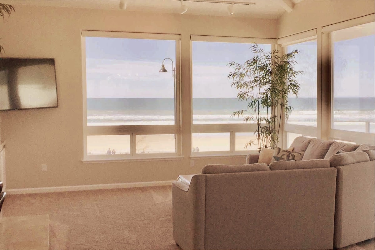 Large windows frame a view of the ocean, allowing natural light to fill the living area. A sectional sofa is positioned for comfort, with a flat-screen TV mounted on the adjacent wall. Light-colored walls and soft decor create a serene atmosphere.