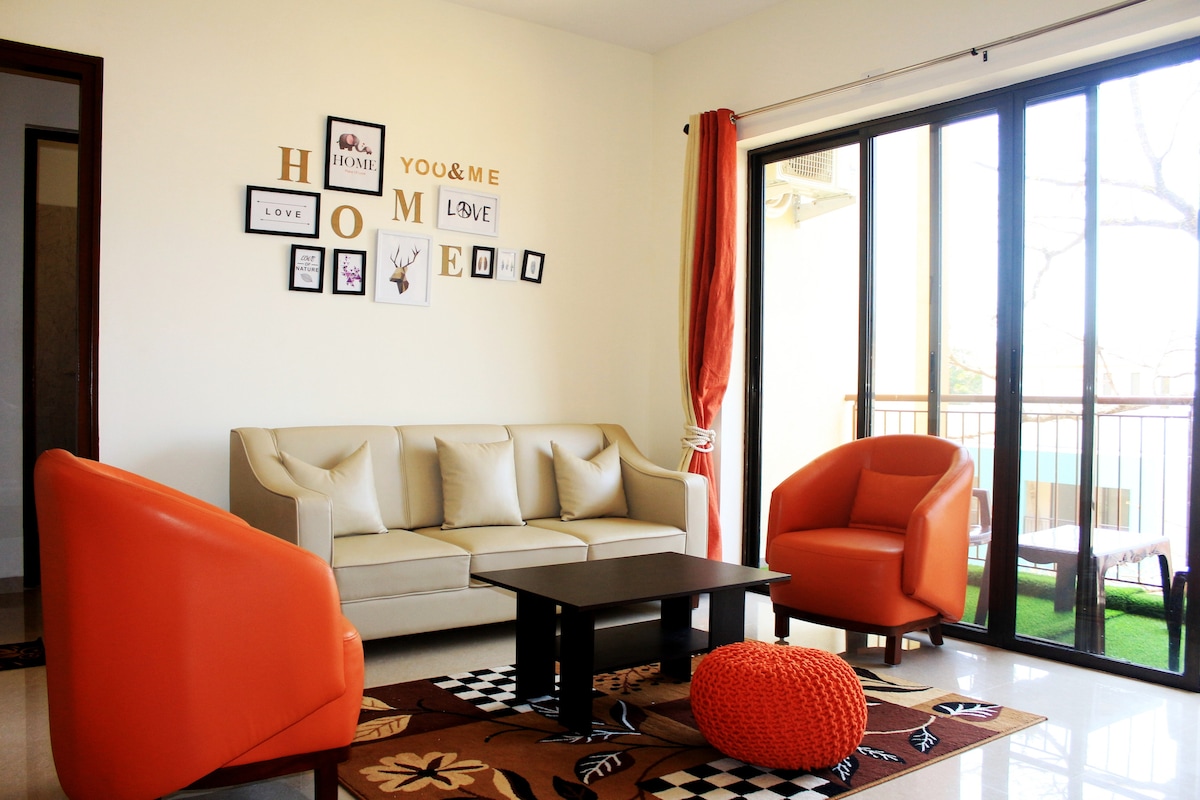 A well-lit living area features a light-colored sofa accompanied by two vibrant orange armchairs. A dark coffee table sits on a decorative rug, while framed pictures adorn the wall above. Large glass doors lead to a balcony, enhancing the airy feel of the space.