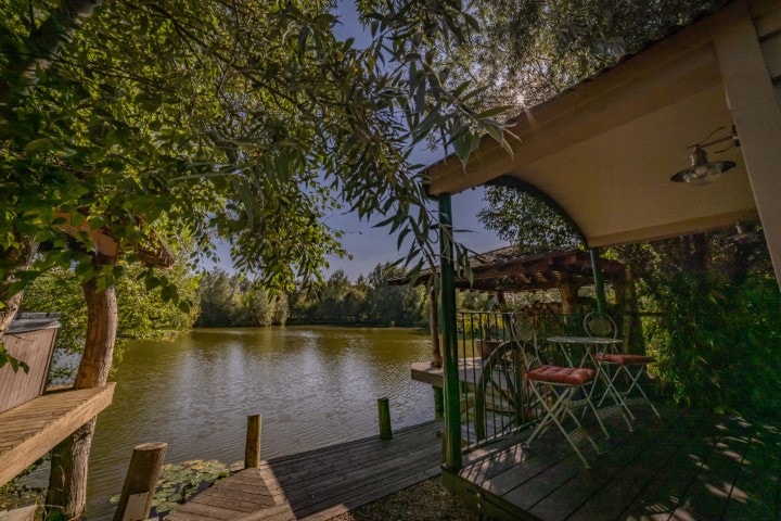 A serene view of the adjacent lake is framed by lush greenery. A covered deck features a small table and chairs, inviting relaxation. The calm water reflects the surrounding trees, creating a peaceful atmosphere.