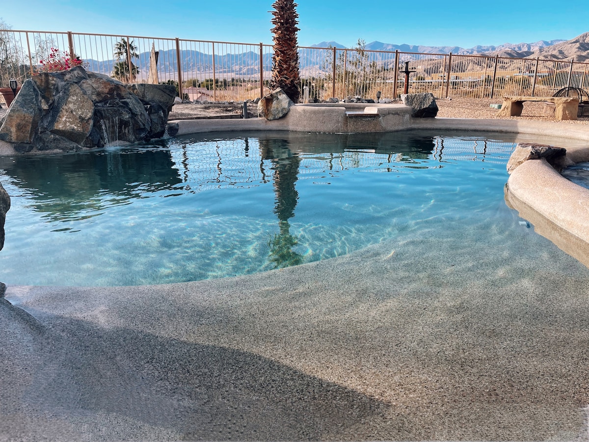 A clear saltwater pool is surrounded by natural stone features, reflecting the sunlight. The edge of the pool gently flows into the expansive outdoor area, with picturesque mountain views visible in the background, adding to the serene atmosphere.