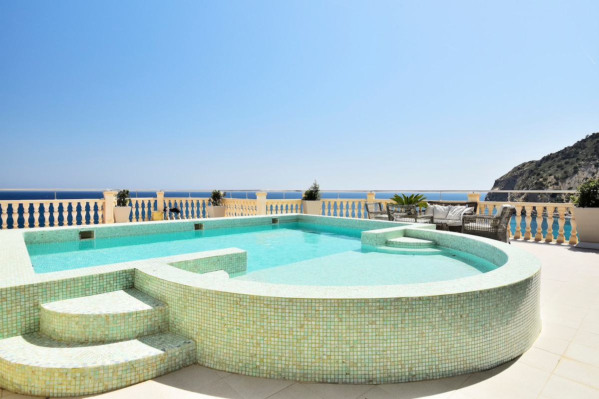 A private swimming pool is displayed with a mosaic tile design, offering a spacious area for relaxation. Clear blue water reflects the sky, and a sun terrace is visible around the pool, surrounded by planters. Views of the ocean and hillside are in the background.