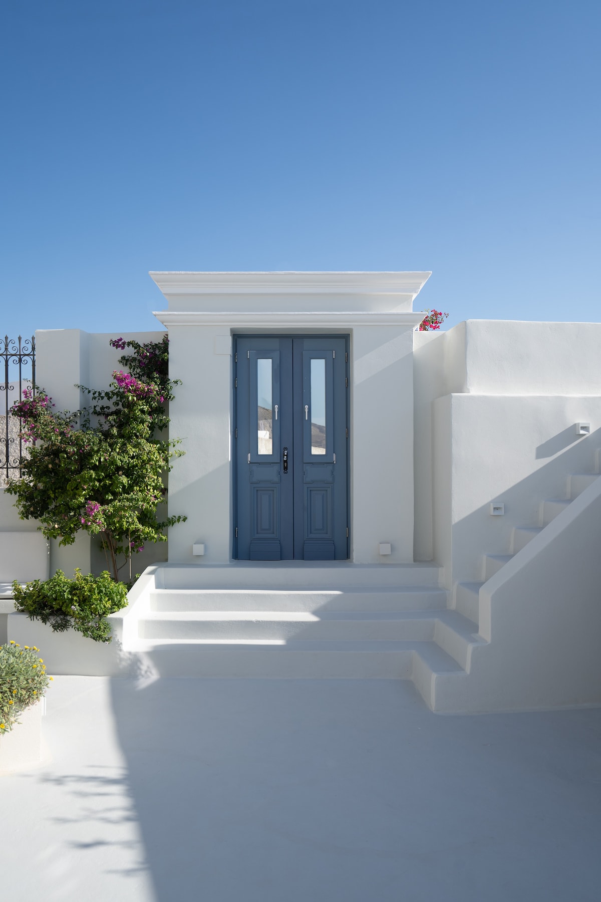 A charming entrance features a pair of blue doors framed by white walls, complemented by vibrant blooming bougainvillea. A staircase leads upwards, bordered by lush greenery against a clear blue sky, creating a serene and inviting approach to the property.