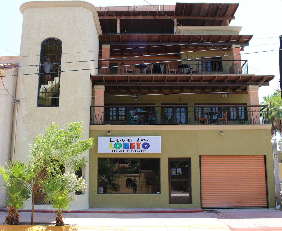 A multi-story building is displayed, featuring a ground-level business with large signage and a roll-up door. Balconies are visible on the upper floors, equipped with chairs. Adjacent greenery adds a vibrant touch, while clear blue skies provide a backdrop.