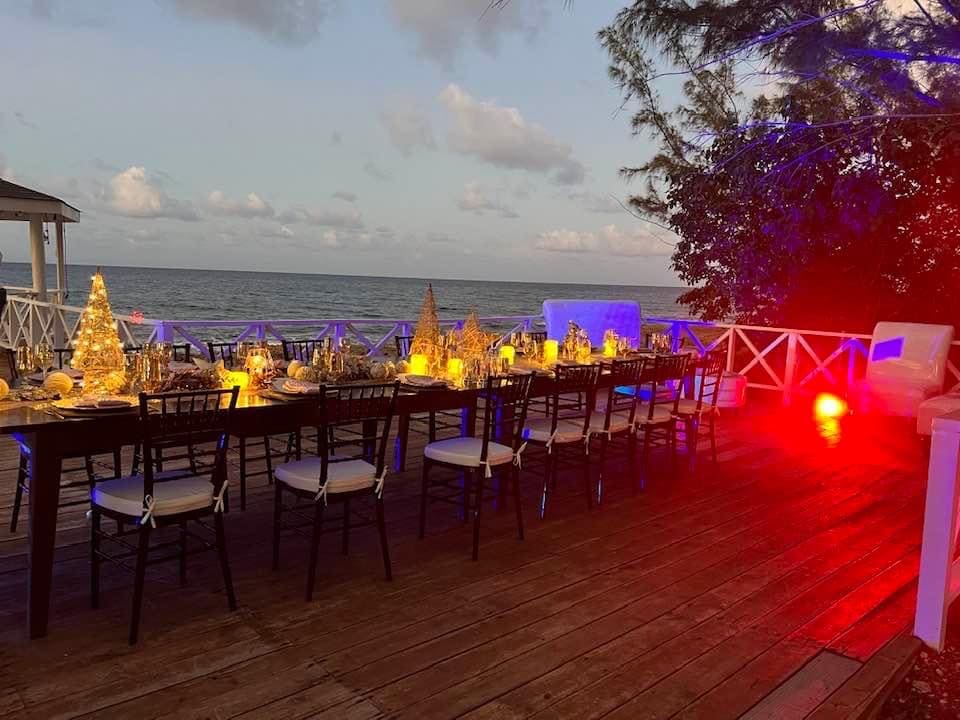 A spacious outdoor dining area is set up on a wooden deck overlooking the ocean. A long table is adorned with elegant place settings and illuminated decorative elements. Soft lighting enhances the inviting ambiance, with the gentle waves visible in the background.