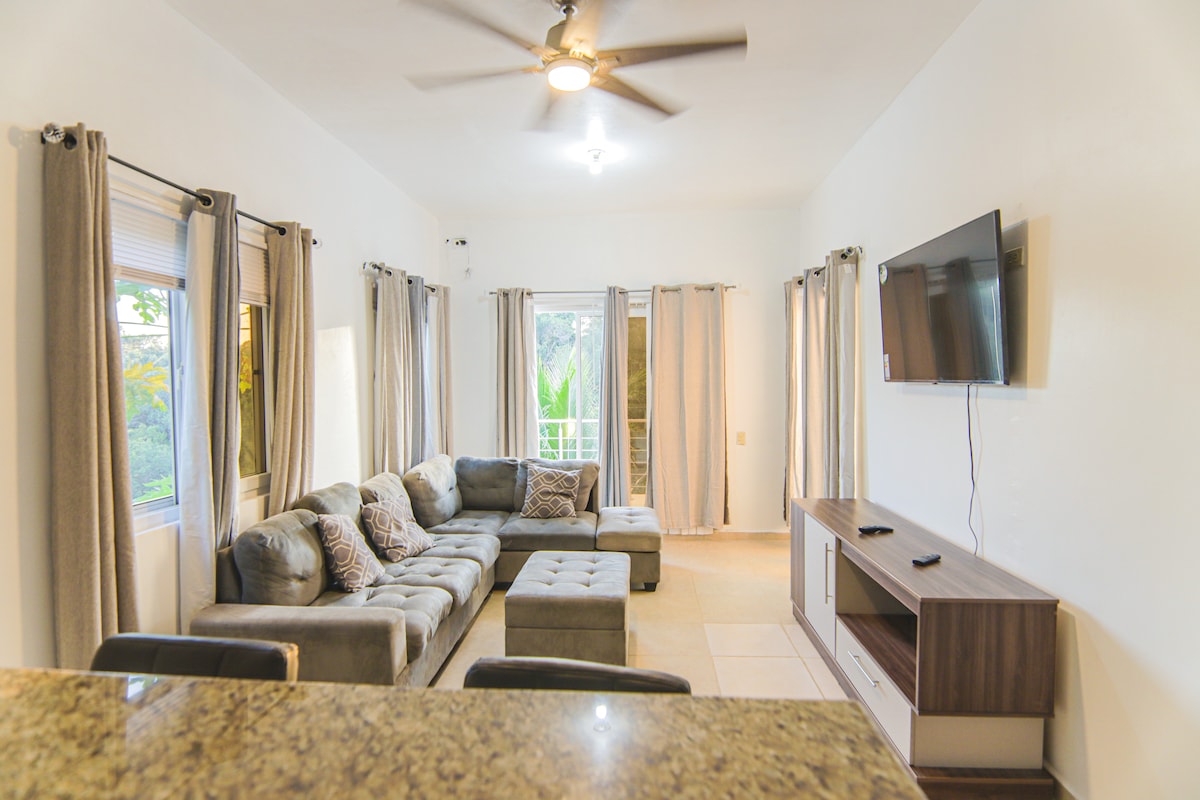 A spacious living room features a large sectional sofa, complemented by several decorative pillows. Natural light filters in through multiple windows adorned with soft curtains. A mounted television is positioned on the wall, and a stylish console table is placed beneath it.