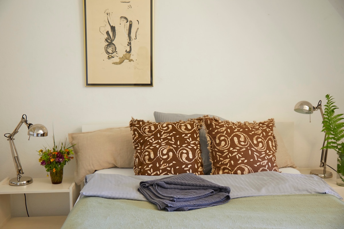 The bedroom features a neatly made bed with decorative brown pillows and a light green throw. Two bedside tables hold modern lamps and a small bouquet of flowers, while a framed artwork adds a subtle touch to the wall above.