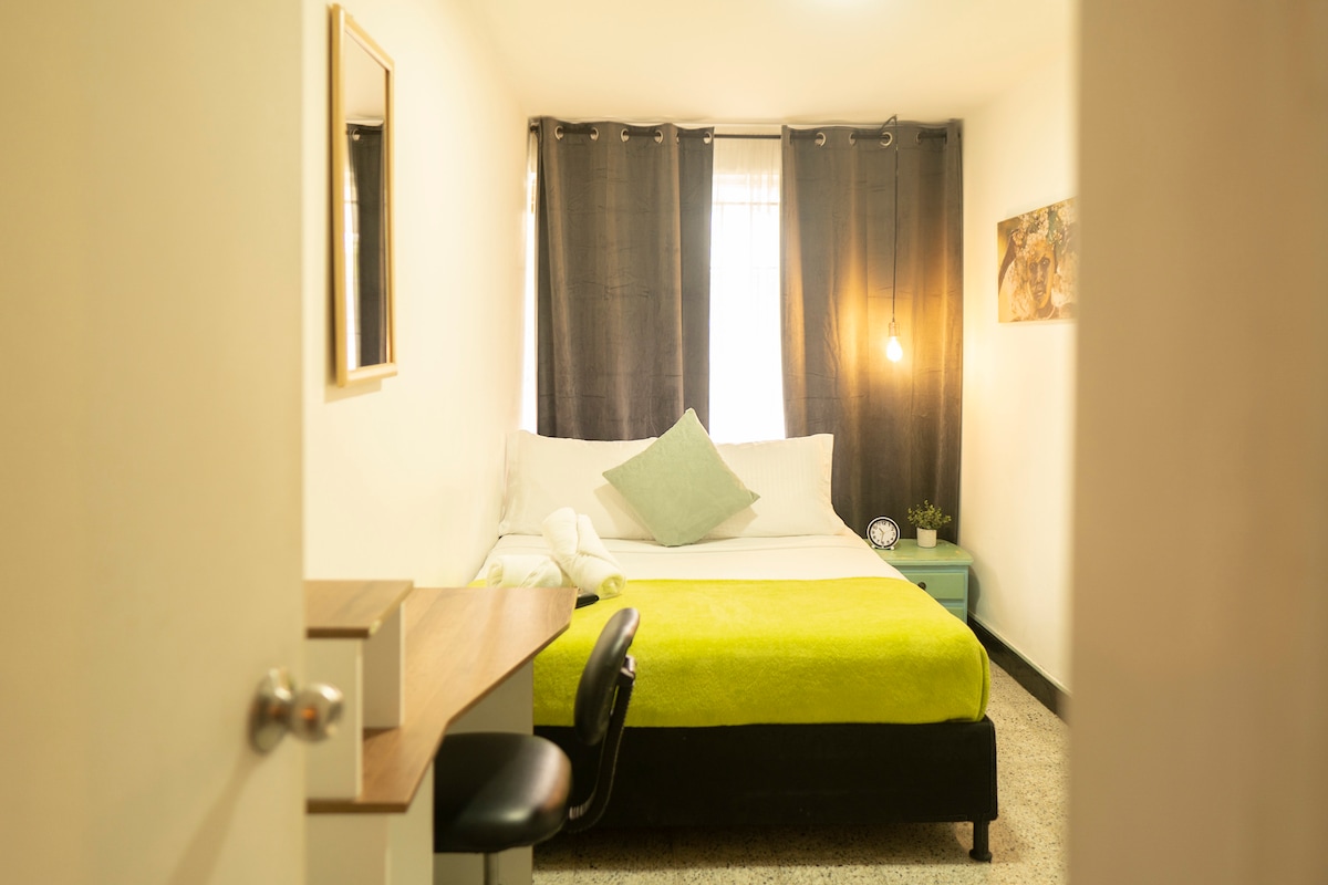 A private room features a spacious bed covered with a green blanket, complemented by a light blue pillow. A desk with a chair is included, and a mirror is mounted on the wall. Natural light enters through large windows, framed by dark curtains.