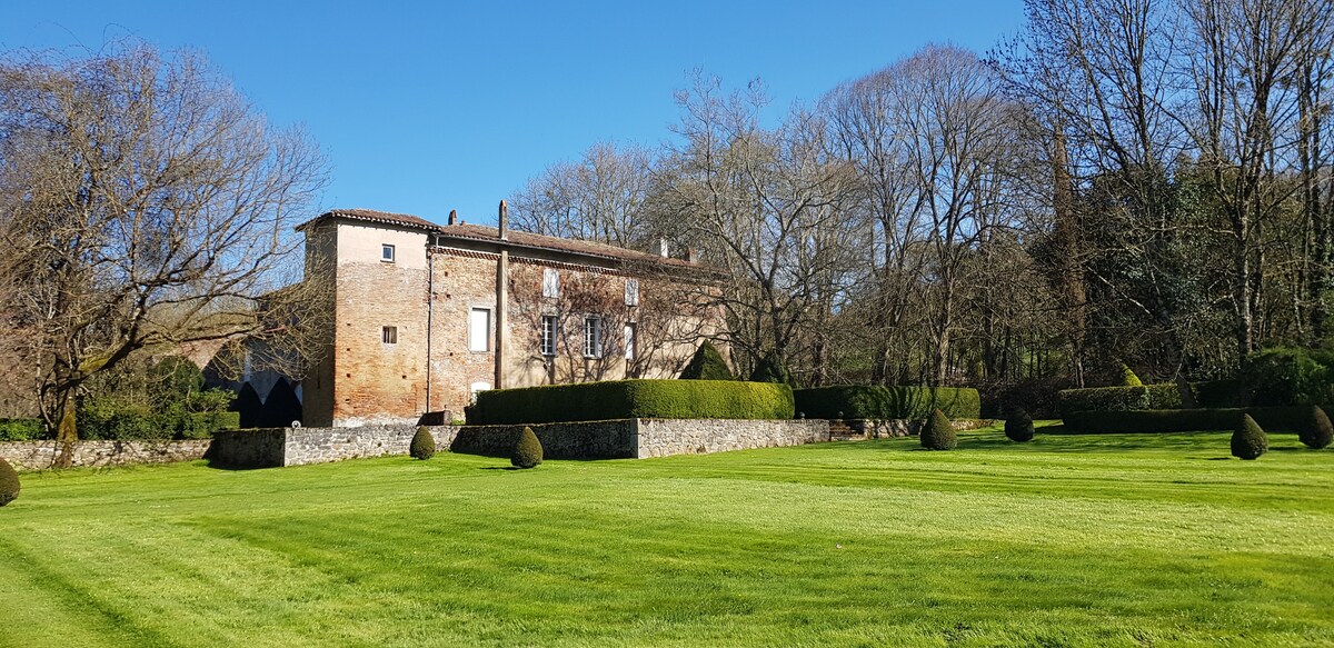 The image captures a spacious green lawn bordered by neatly trimmed hedges and trees. A two-story brick building with a simple facade stands in the background, complementing the clear blue sky above. The serene landscape invites relaxation amidst nature.