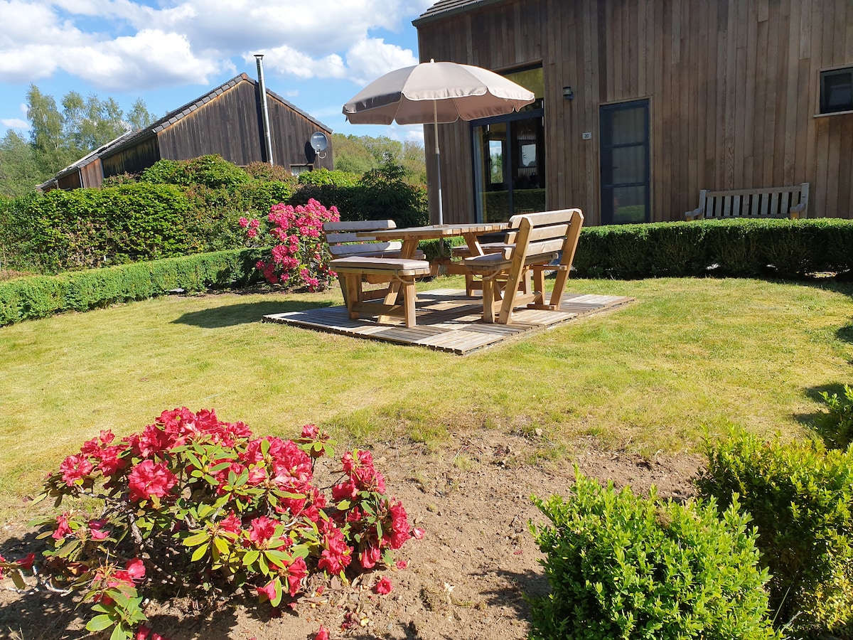 A landscaped outdoor area features a wooden dining table surrounded by four benches, shaded by a large umbrella. Colorful flowering bushes add vibrancy to the space, complemented by manicured grass. Nearby, a wooden bench provides an additional seating option.