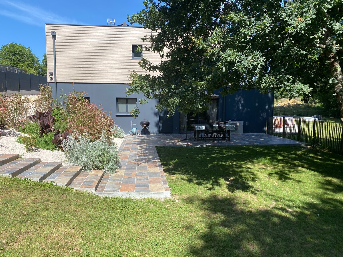 The image shows a spacious outdoor area with a well-maintained garden and patio set. The property features a combination of stone pathways and grass, surrounded by shrubbery and trees. A barbecue grill is visible near the dining table, offering an inviting space for outdoor gatherings.