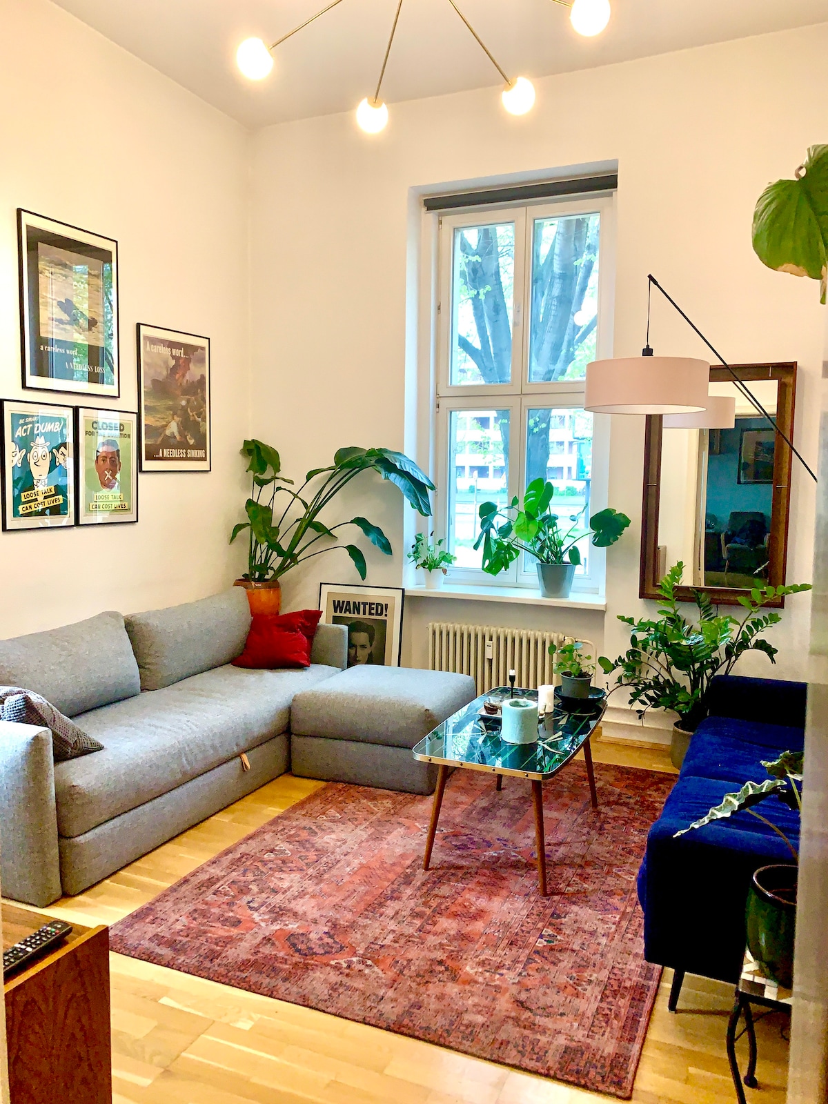 The living room features a gray sectional sofa arranged beside a blue velvet couch. A glass coffee table is positioned centrally, adorned with plants and cups. Bright green plants add vibrancy, while framed artwork decorates the walls. A large window allows natural light to fill the space.