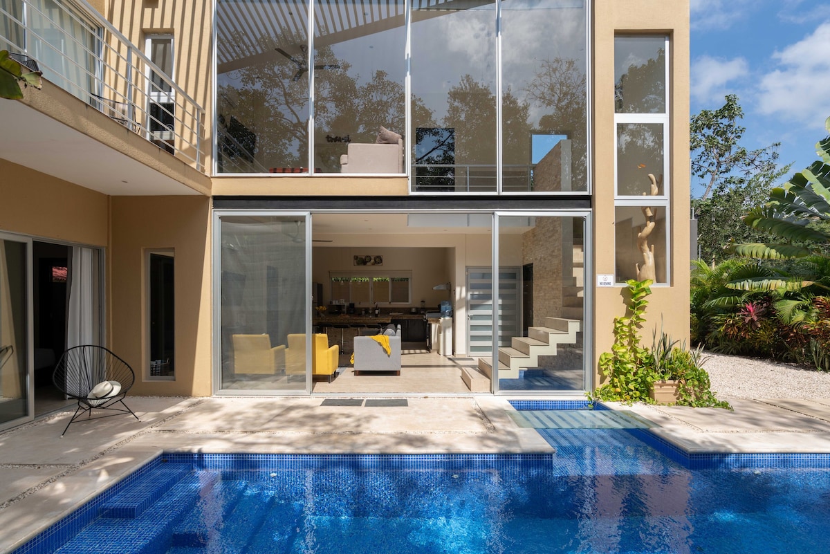A modern home is set beside a private pool, featuring expansive glass walls that invite natural light. Lush greenery surrounds the area, complementing the outdoor seating arrangements. Clear water reflects the open sky, creating a serene atmosphere for relaxation.
