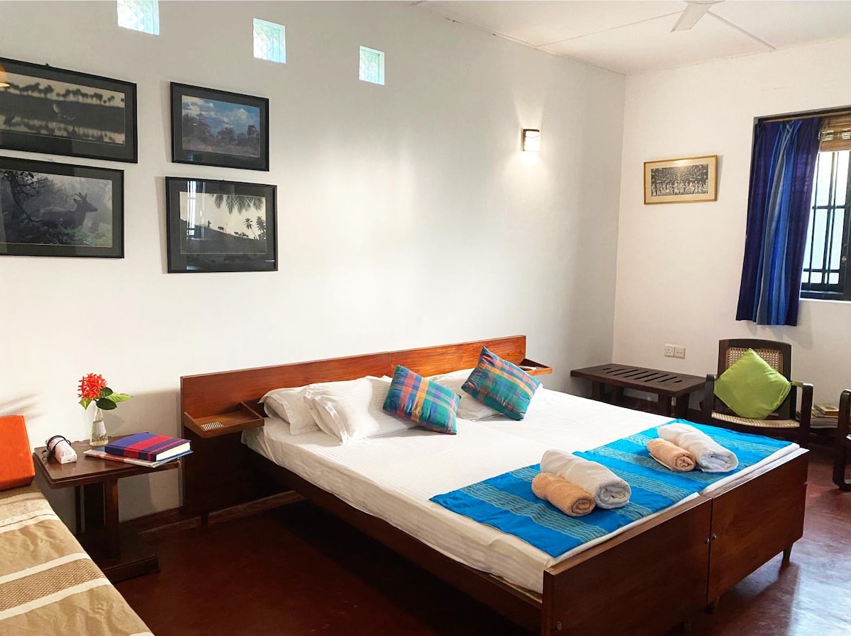 A spacious room is furnished with two beds, decorative wall art, and a vibrant blue throw. A small table and comfortable chairs provide a relaxing space, while natural light enters through a nearby window. Fresh towels are neatly arranged on the beds.