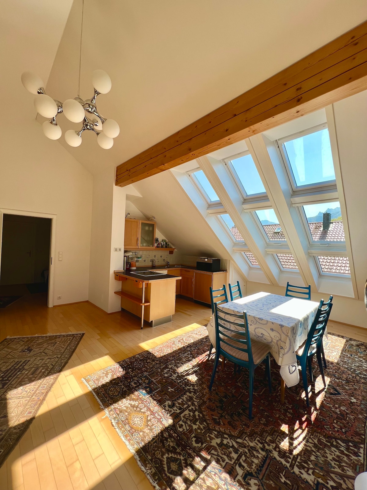 An airy dining area is highlighted by a large, light-filled window with a sloped ceiling. A rectangular table with seating for six is positioned on a patterned rug, surrounded by blue chairs. The kitchen area is visible to the side, featuring wooden cabinetry.