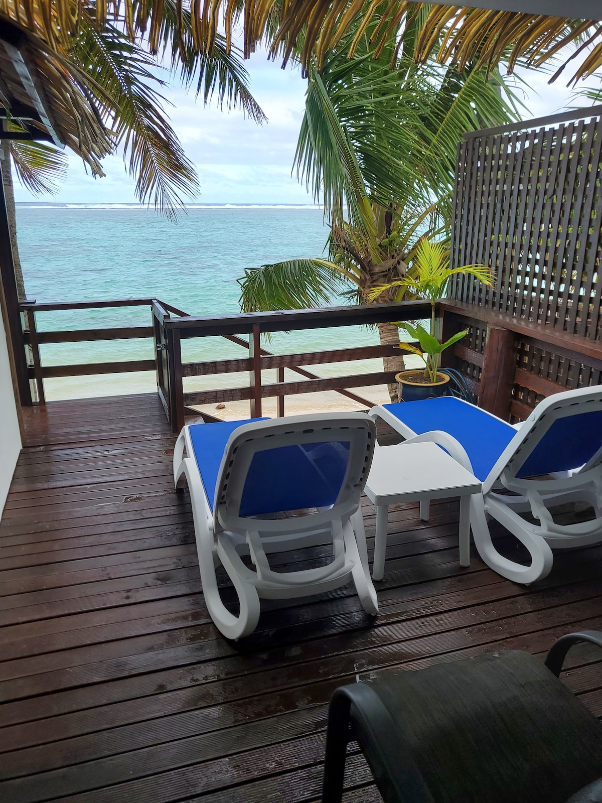 A private deck area is presented with two blue loungers and a small white table, surrounded by tropical plants. The deck overlooks a serene lagoon, with gentle waves lapping at the shore, framed by swaying palm trees and a wooden railing.