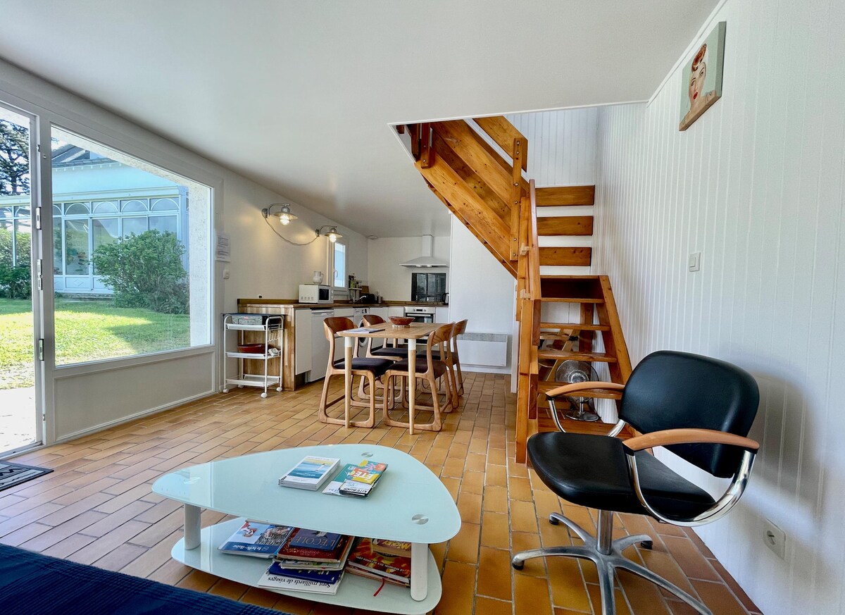 A spacious living area is illuminated by natural light streaming through large windows. The space features a wooden staircase leading to the upper level, a dining table with chairs, and a stylish black chair. A coffee table holds various magazines, completing the inviting atmosphere.