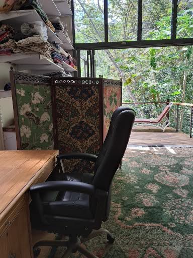 A workspace features a wooden desk and an office chair positioned in front of a colorful folding screen. Natural light is seen streaming in from large glass doors, which open to a deck surrounded by lush greenery, including native plants and trees.