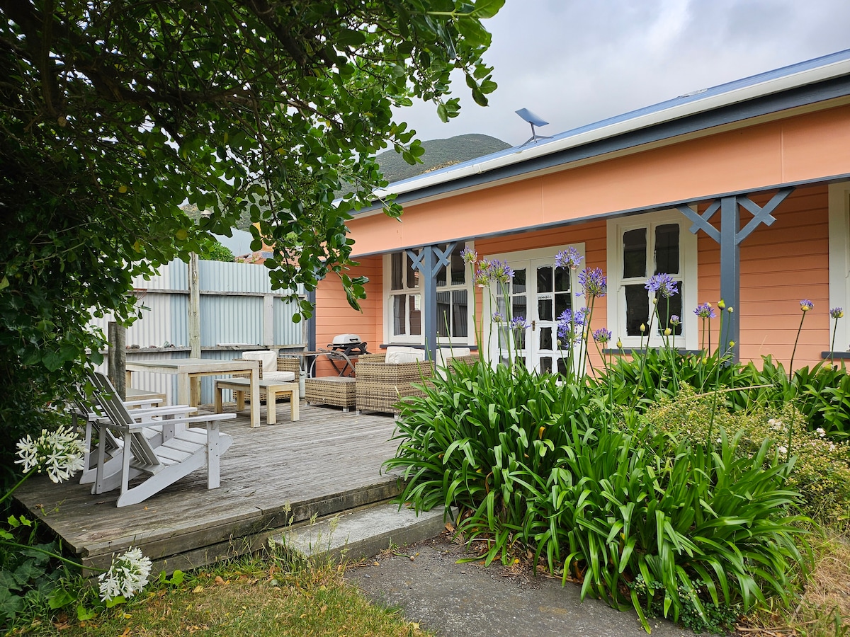 A deck area features outdoor seating arrangements, surrounded by lush green foliage and flowering plants. The cottage's exterior is painted in soft peach tones, with large windows allowing natural light to fill the space. A barbecue is positioned to the side for outdoor cooking.