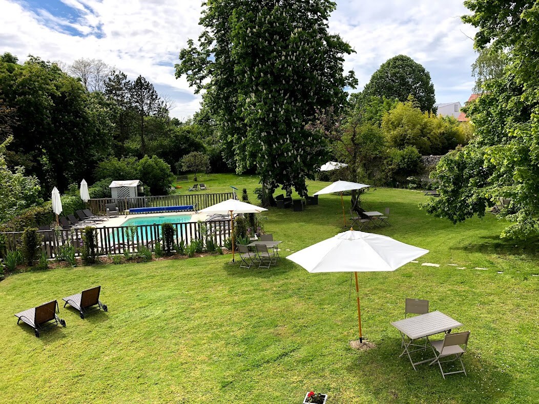 A spacious garden area features a sparkling pool surrounded by lounge chairs and umbrellas. Lush greenery and trees provide shade and tranquility. Several seating arrangements can be seen throughout the lawn, creating inviting spots for relaxation.