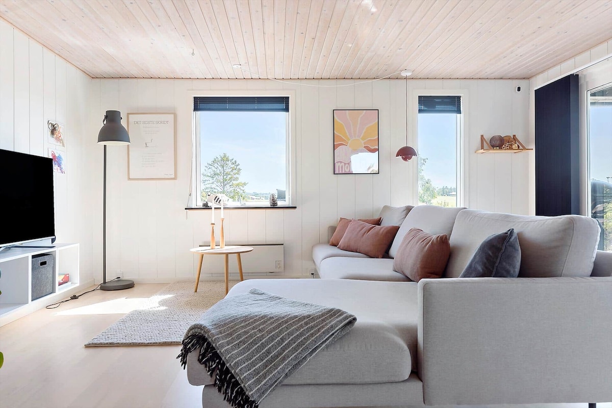A modern living room is presented with large windows allowing natural light to fill the space. A grey sectional sofa is complemented by accent pillows, while a cozy area rug anchors the seating. A television is positioned on a low cabinet, and a floor lamp enhances the ambiance.
