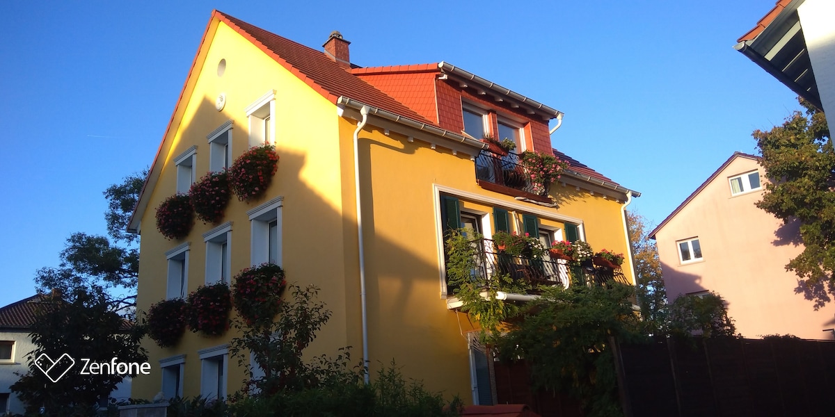 A charming yellow house is presented, characterized by large windows adorned with flower boxes. A balcony is visible on the upper floor, featuring greenery. The clear blue sky serves as a backdrop, enhancing the inviting appearance of the home.