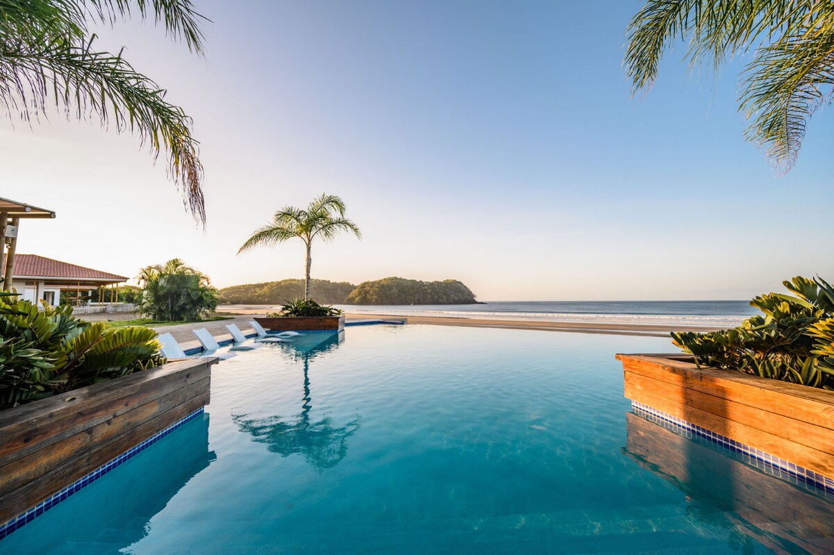 An infinity pool stretches towards the ocean, surrounded by lush greenery and palm trees. The water reflects the soft hues of the sky, creating a serene atmosphere. In the distance, a gentle curve of the beach meets the horizon, inviting relaxation and leisure.