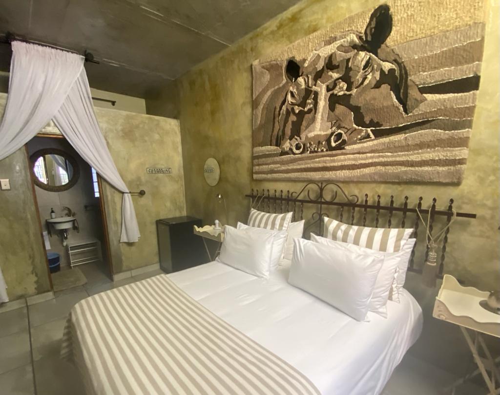 A spacious sleeping area is furnished with two white pillows atop a striped blanket. A woven wall art piece displays intricate designs. A private bathroom entrance is visible, and a small fridge is positioned near the wall, adding convenience to the room's layout.