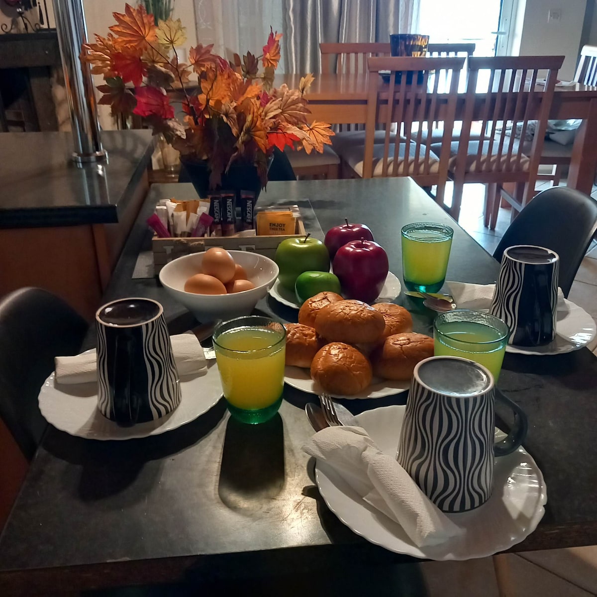 A dining table is set with a variety of breakfast items, including eggs, fruits, and pastries. Colorful cups and plates complement the arrangement. A floral centerpiece adds a touch of nature, while natural light highlights the inviting atmosphere.