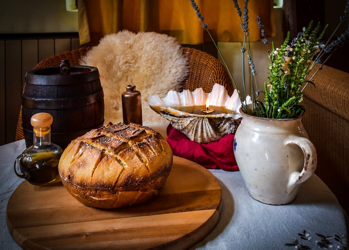 A rustic table is set with a round loaf of freshly baked bread on a wooden board. An olive oil bottle and a spice grinder are placed nearby. A ceramic pitcher and a small vase containing fresh herbs complete the arrangement, creating a warm, homey setting.