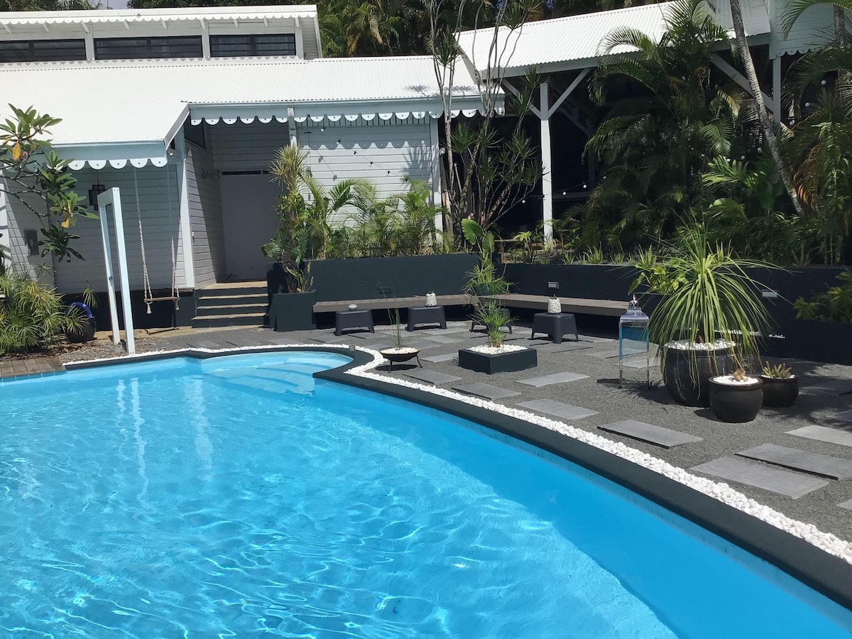 A clean swimming pool glistens under the sun, surrounded by landscaped greenery and several lounge chairs positioned along the edge. The light-colored wooden lodge is visible in the background, offering shaded steps leading up to a larger deck area.