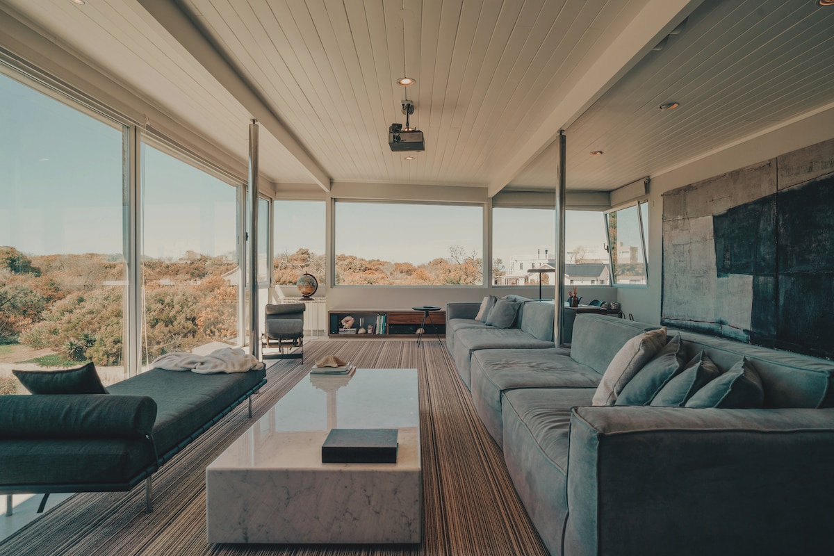A modern living area features large windows offering views of the surrounding natural landscape. A spacious sectional sofa and lounge chair provide comfortable seating. A minimalist coffee table stands at the center, and light-colored wood flooring complements the design.
