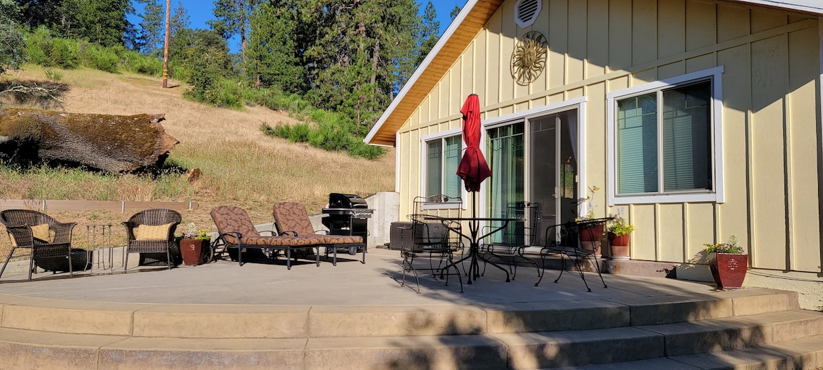 A spacious patio features a seating area with several comfortable chairs and a table, surrounded by plants. A barbecue grill is positioned nearby. The backdrop consists of a gently sloping hillside with trees, contributing to the peaceful outdoor setting.