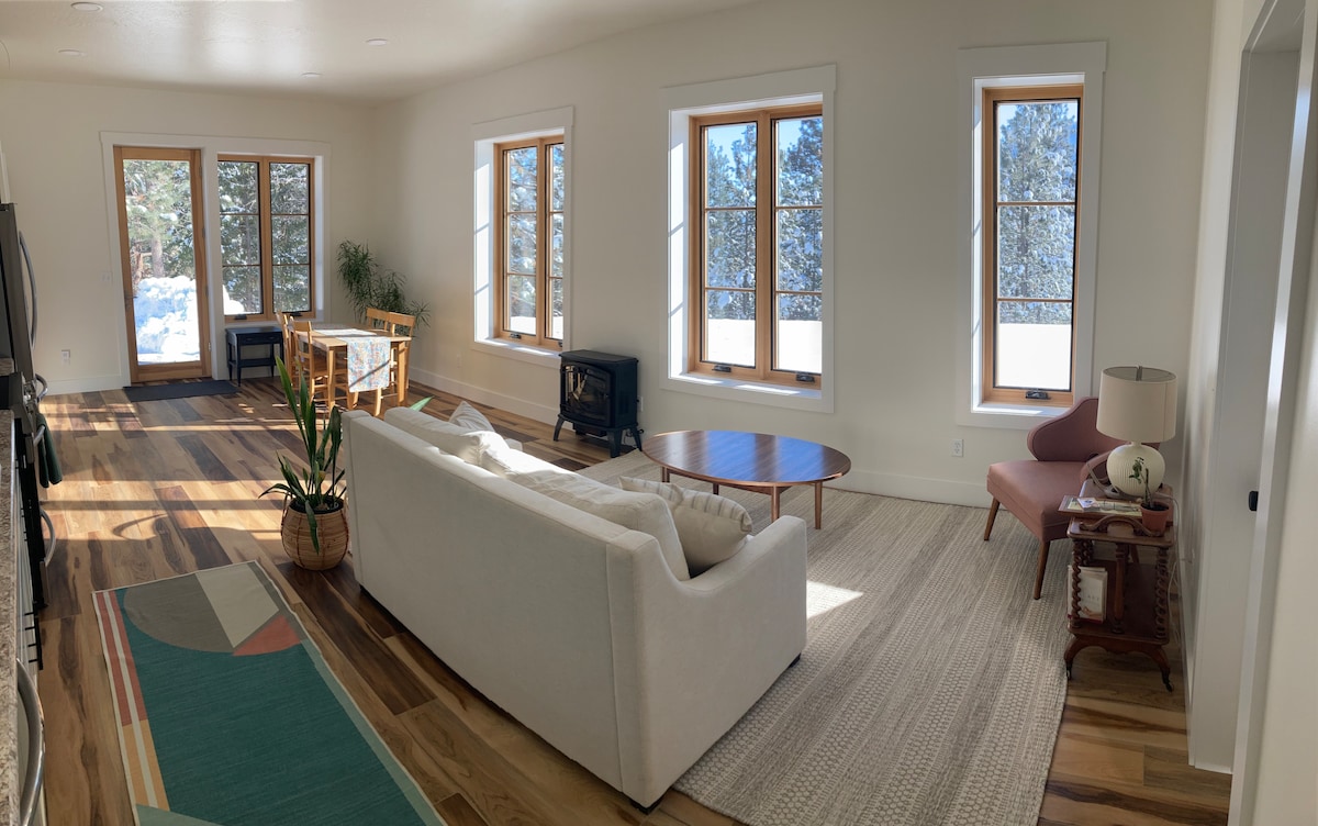 A spacious living area features large windows that fill the space with natural light. A comfortable sofa faces a coffee table, while a small dining table is positioned nearby. A plant adds a touch of greenery to the neutral decor.