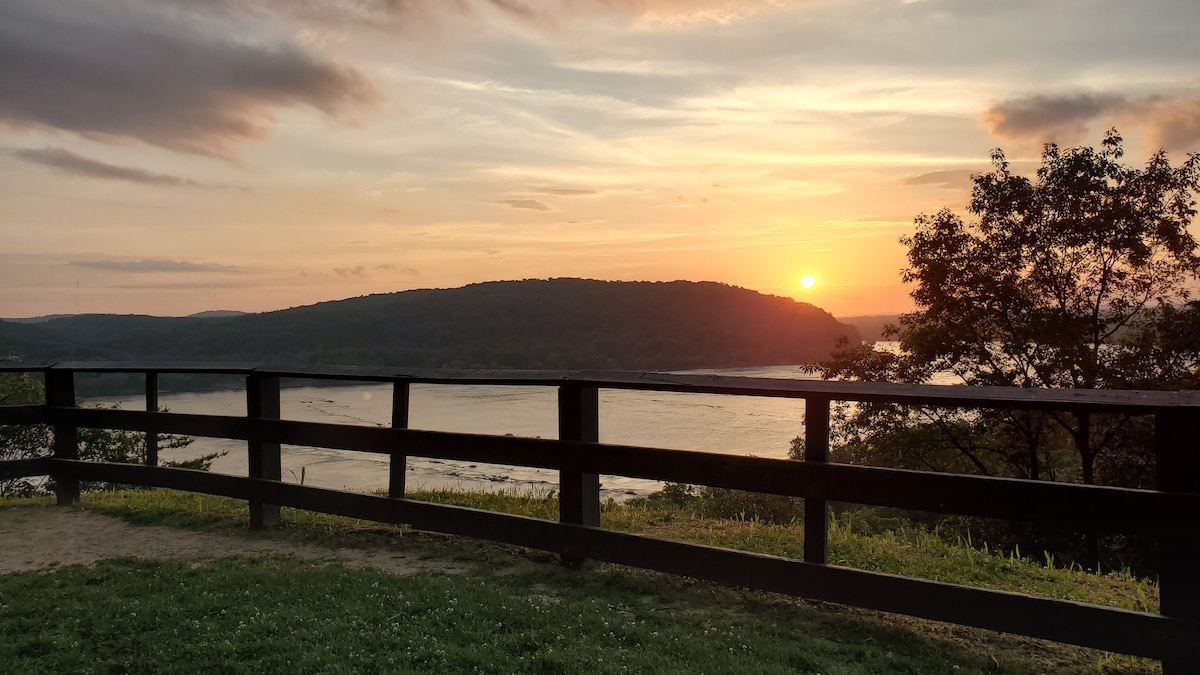A wooden railing frames a scenic overlook with a vibrant sunset casting warm hues over the Susquehanna River. A distant hill is visible across the water, while trees and grassy areas surround the viewpoint, providing a peaceful natural setting.