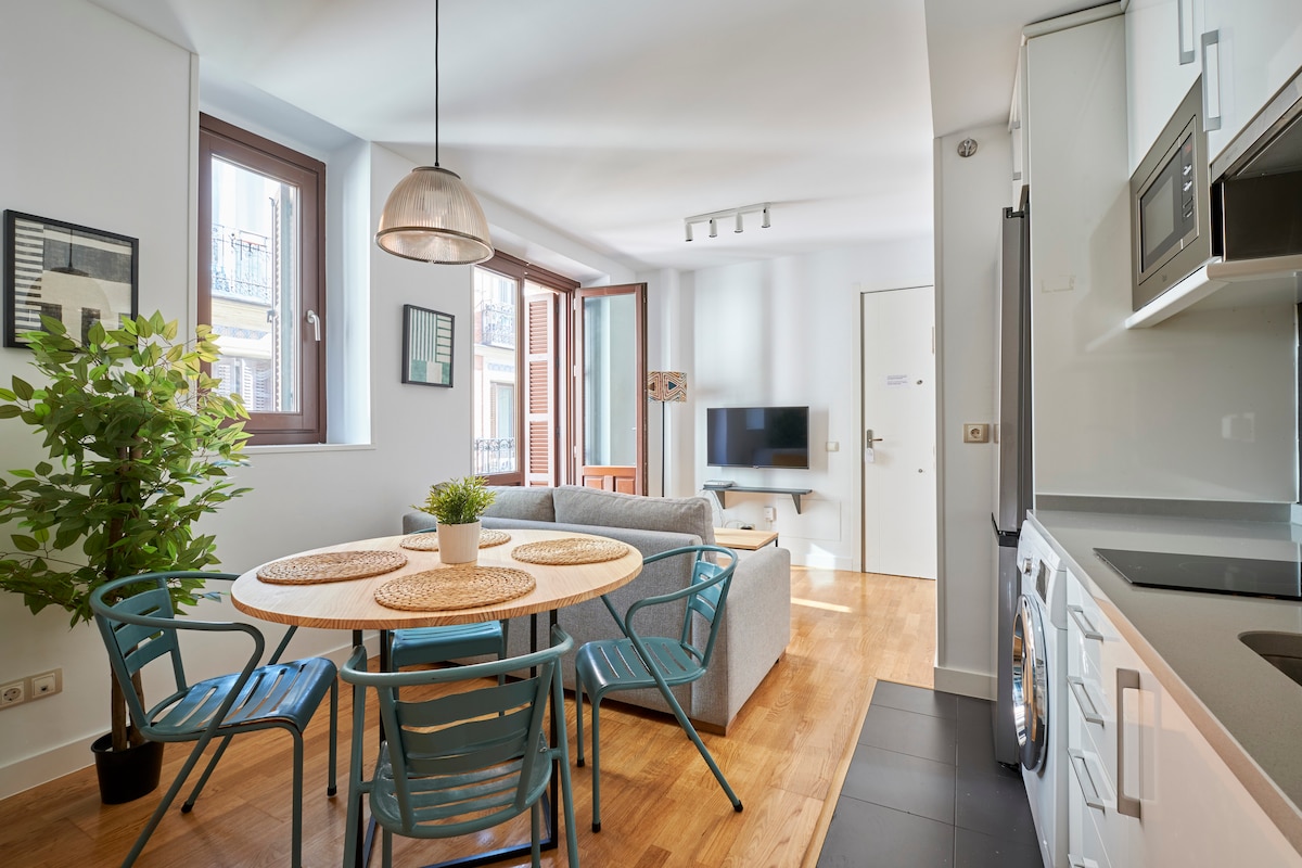 A bright dining area features a round table with woven placemats surrounded by blue chairs. Sunlight pours in through large windows, illuminating the adjacent living space, which includes a gray sofa and a television. Potted greenery adds a touch of nature.