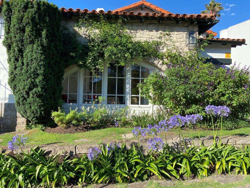 The exterior of the property features a charming stone façade, framed by flourishing green foliage. Colorful purple flowers line the well-maintained garden beds, contributing to the inviting appearance of the home. Large windows allow natural light to illuminate the interiors.