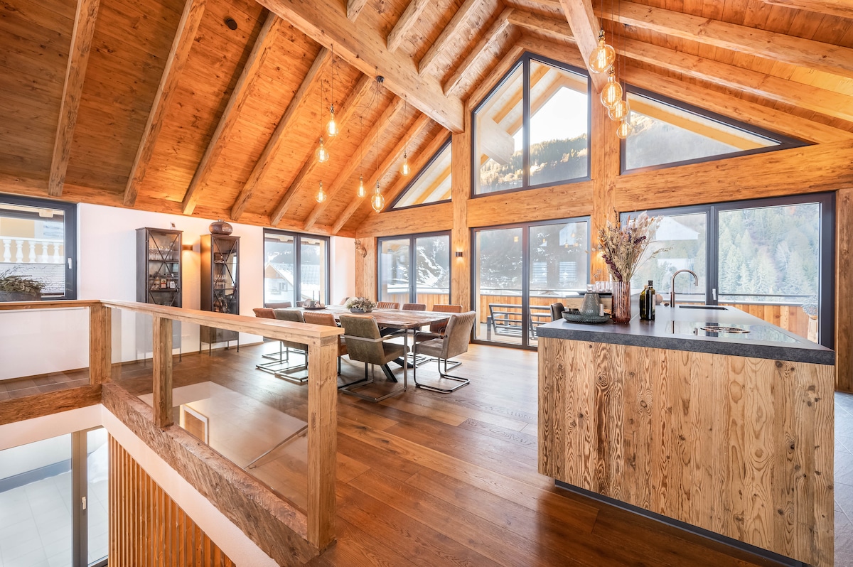 An open-plan living area features a high wooden beam ceiling with large windows showcasing mountain views. A stylish kitchen island is positioned adjacent to a large dining table, surrounded by comfortable seating. Natural wood elements on the floor and furnishings add warmth to the space.