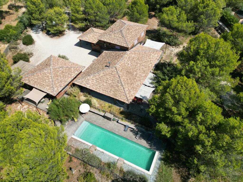 An aerial view of a wooden house complex surrounded by lush greenery, featuring multiple rooftops with terracotta tiles. A serene swimming pool is visible, bordered by a stone patio, with shaded seating areas nearby.