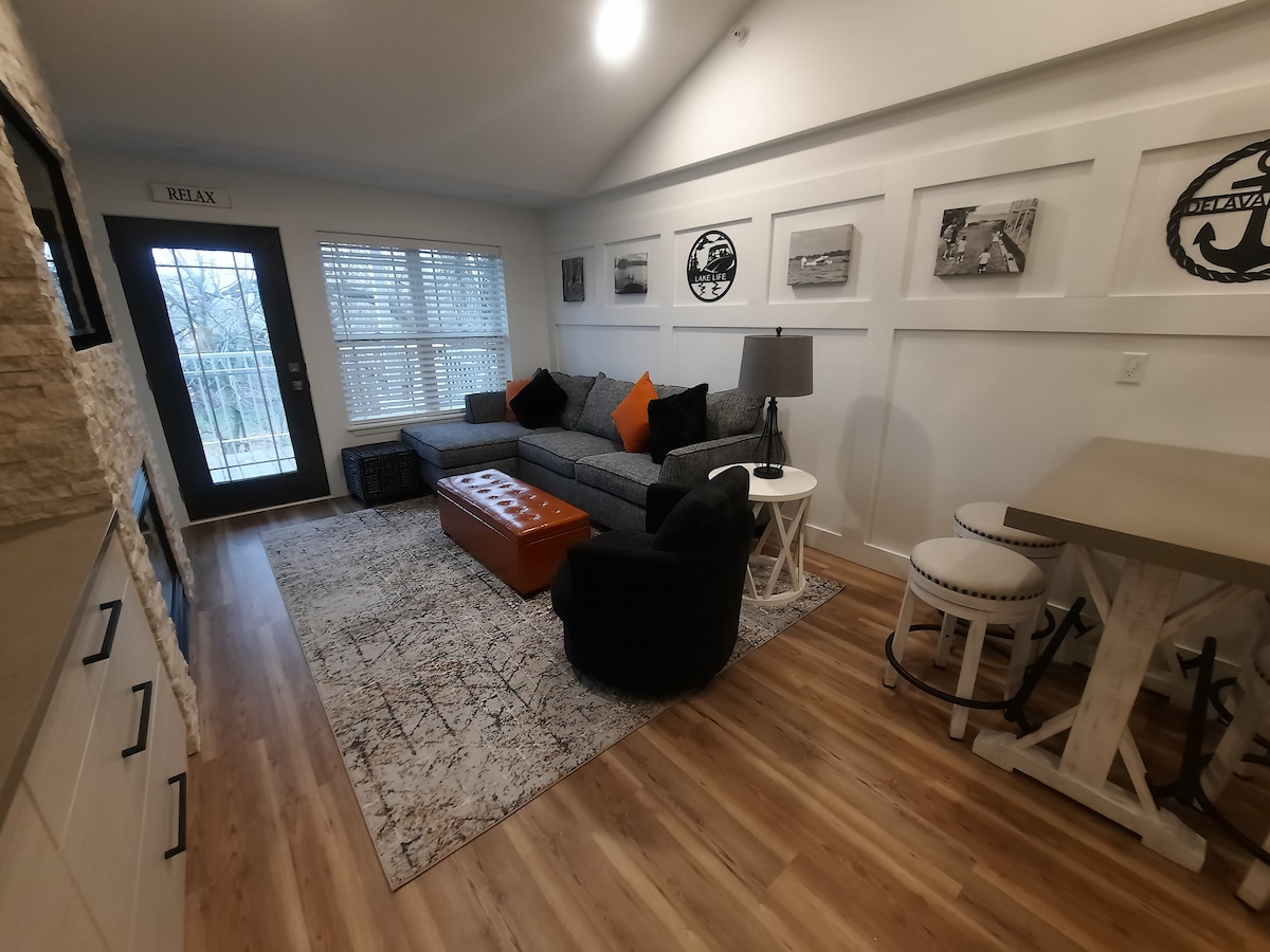 A comfortable living area is highlighted by a spacious sectional sofa adorned with orange accent pillows. A wooden coffee table sits in front, complemented by a patterned area rug. Natural light filters through large windows, and a small dining table with stools is visible, enhancing the inviting space.