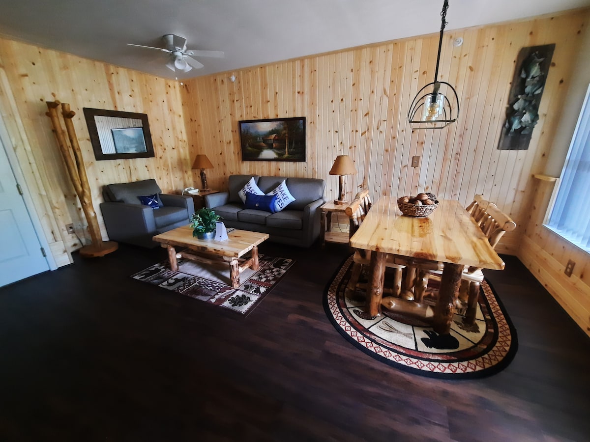 A cozy living space features warm wooden walls and dark flooring. Two comfortable sofas are positioned beside a rustic coffee table. A wooden dining table surrounded by chairs occupies one corner, complemented by decorative items. Natural light filters through the nearby window.