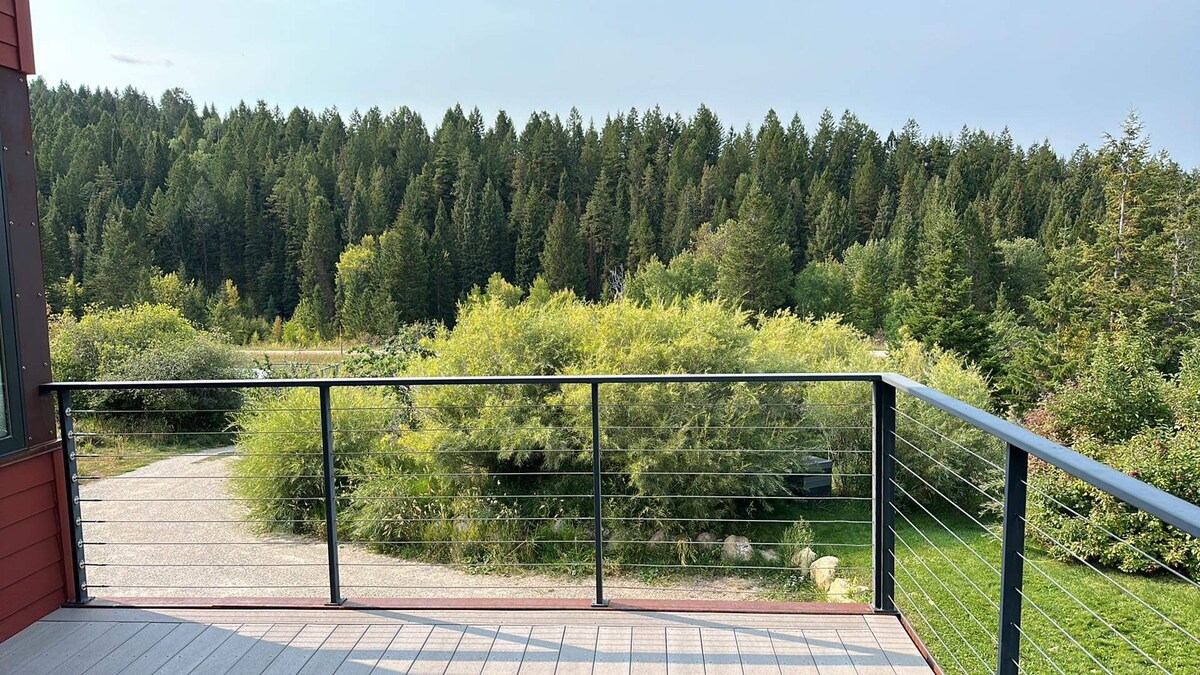 An elevated private deck features a railing with a wire design, overlooking a serene green landscape. Trees and shrubs fill the foreground, creating a natural buffer that enhances the peaceful setting. The distant forest stretches across the horizon under a clear sky.
