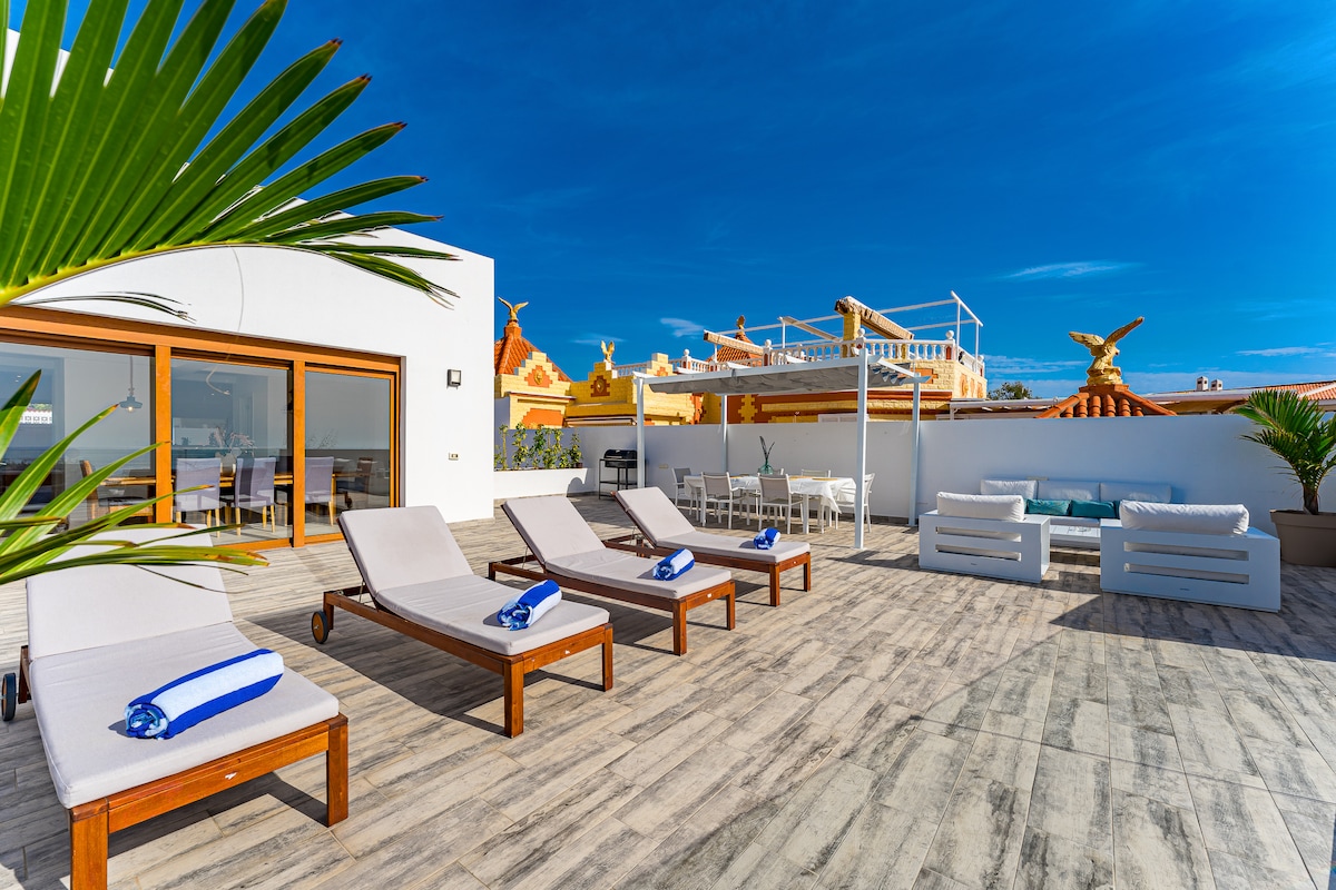 A spacious terrace is highlighted by four sun loungers, neatly arranged and adorned with blue towels. Light wooden flooring complements the vibrant blue sky above. In the background, a dining area and a lounge space are visible, surrounded by minimalist decor and lush greenery.