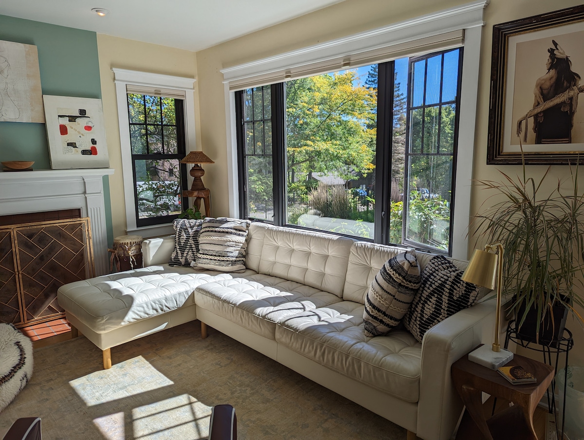 A spacious living area features a white sectional sofa that provides ample seating. Large windows create a bright ambiance while framing views of the vibrant greenery outside. Decorative pillows and a side table add functional charm to the comfortable space.
