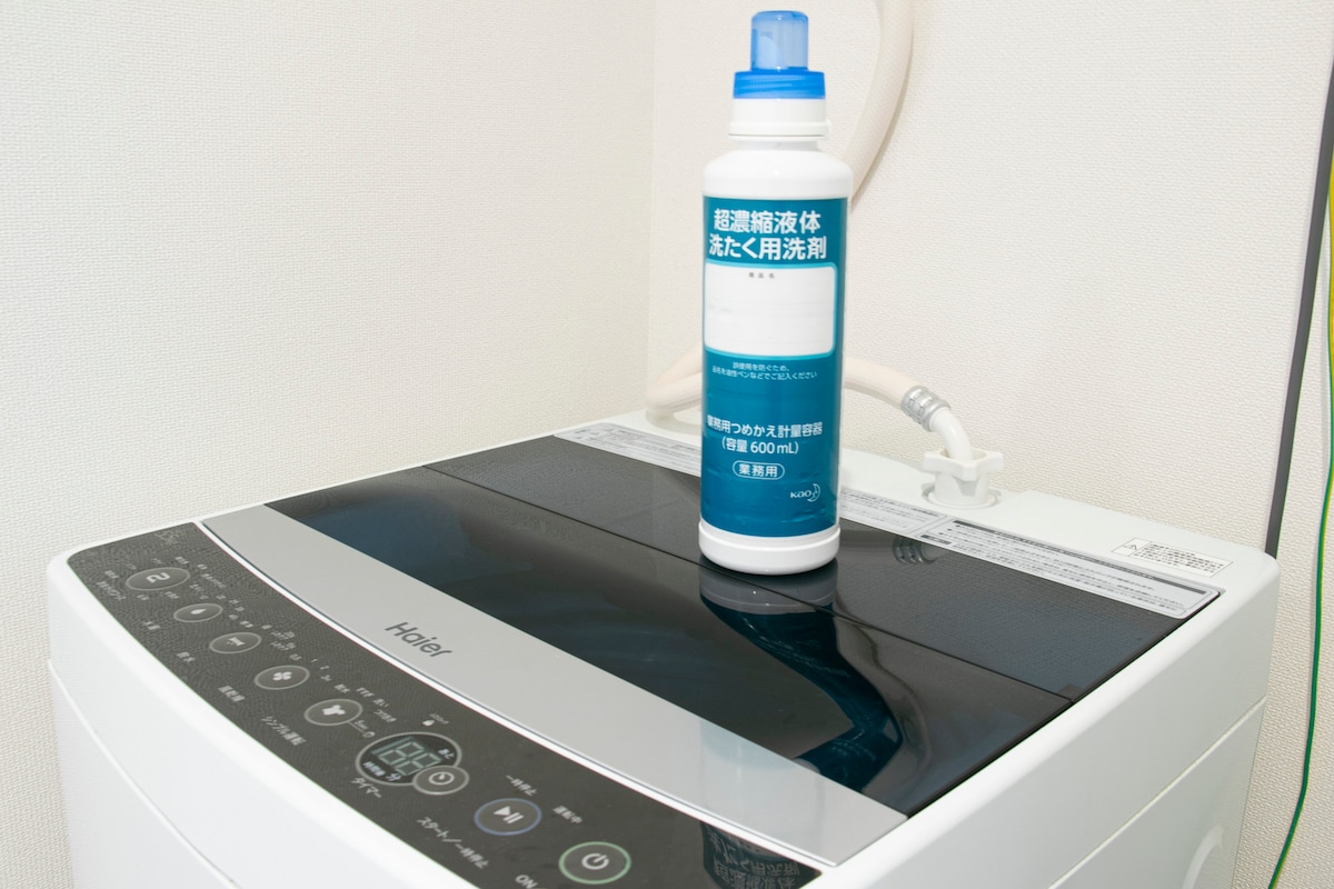 A washing machine is shown on a clean, white countertop. A bottle of laundry detergent rests on top, labeled clearly for easy identification. The machine has an array of buttons and controls, designed for user convenience.