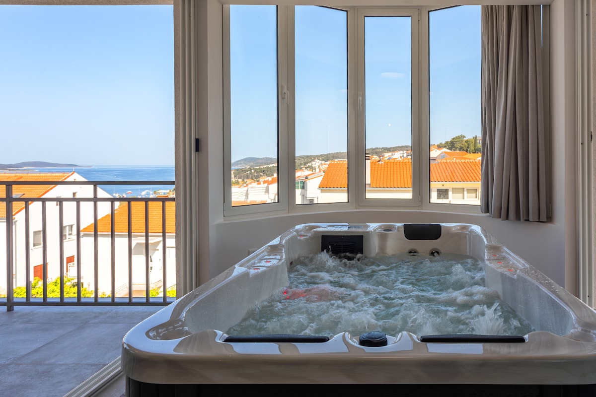 An indoor jacuzzi is positioned near expansive glass windows, offering a view of the sea and surrounding landscape. Natural light fills the space, enhancing the serene atmosphere. Soft curtains frame the windows, complementing the modern design.